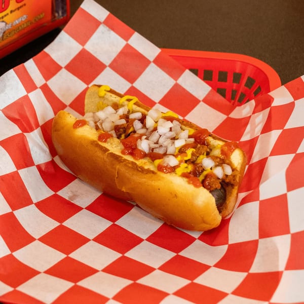 Romeo's Vegan Burgers' chili dog with mustard, ketchup, and diced onions on checkered paper.