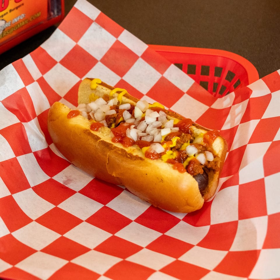 Romeo's Vegan Burgers' chili dog with mustard, ketchup, and diced onions on checkered paper.